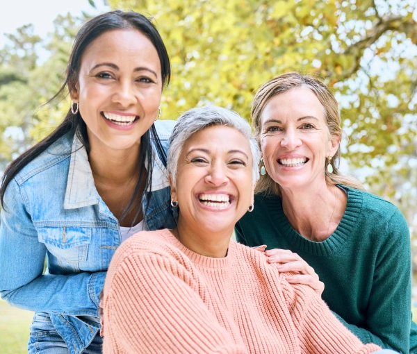 Three smiling women posing together outdoors with yellow-green foliage in background.