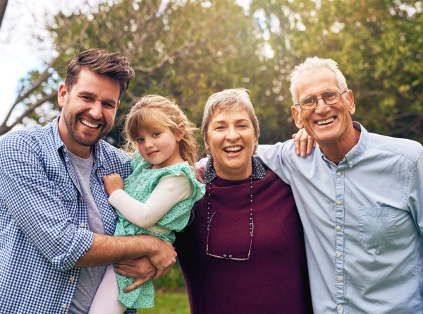 Four generations of a family smiling together outdoors in a park setting.