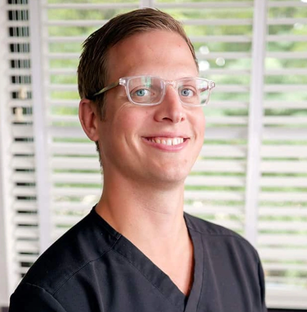 Male dental professional wearing glasses and black scrubs, smiling in clinical office setting.