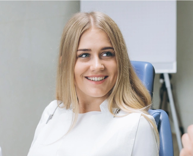 Woman smiling at camera in dental office wearing white shirt, seated in blue chair.