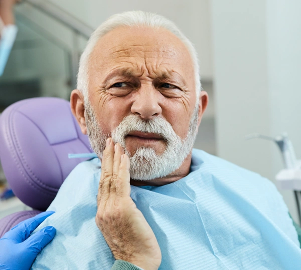 Senior man in dental chair experiencing tooth pain, holding cheek during examination.