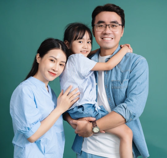 Happy Asian family of three smiling together against turquoise background in casual blue clothing.
