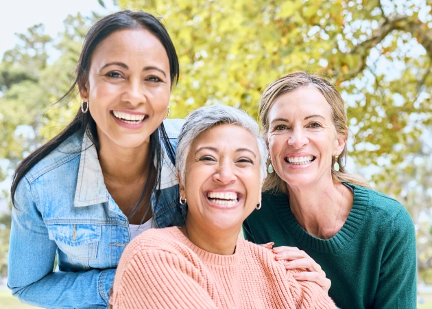Three smiling women posing together outdoors with green foliage background for dental practice portrait.