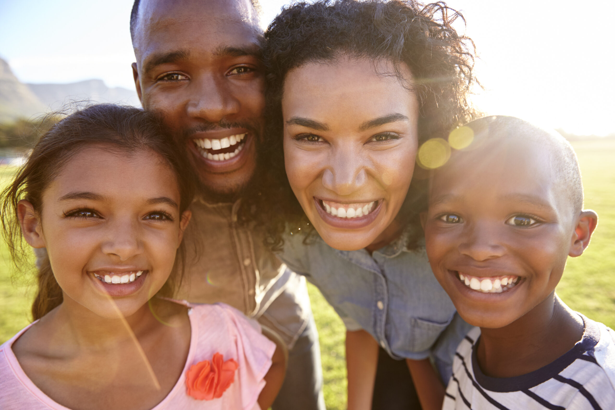 Four smiling children and an adult posing together outdoors in sunny daylight.