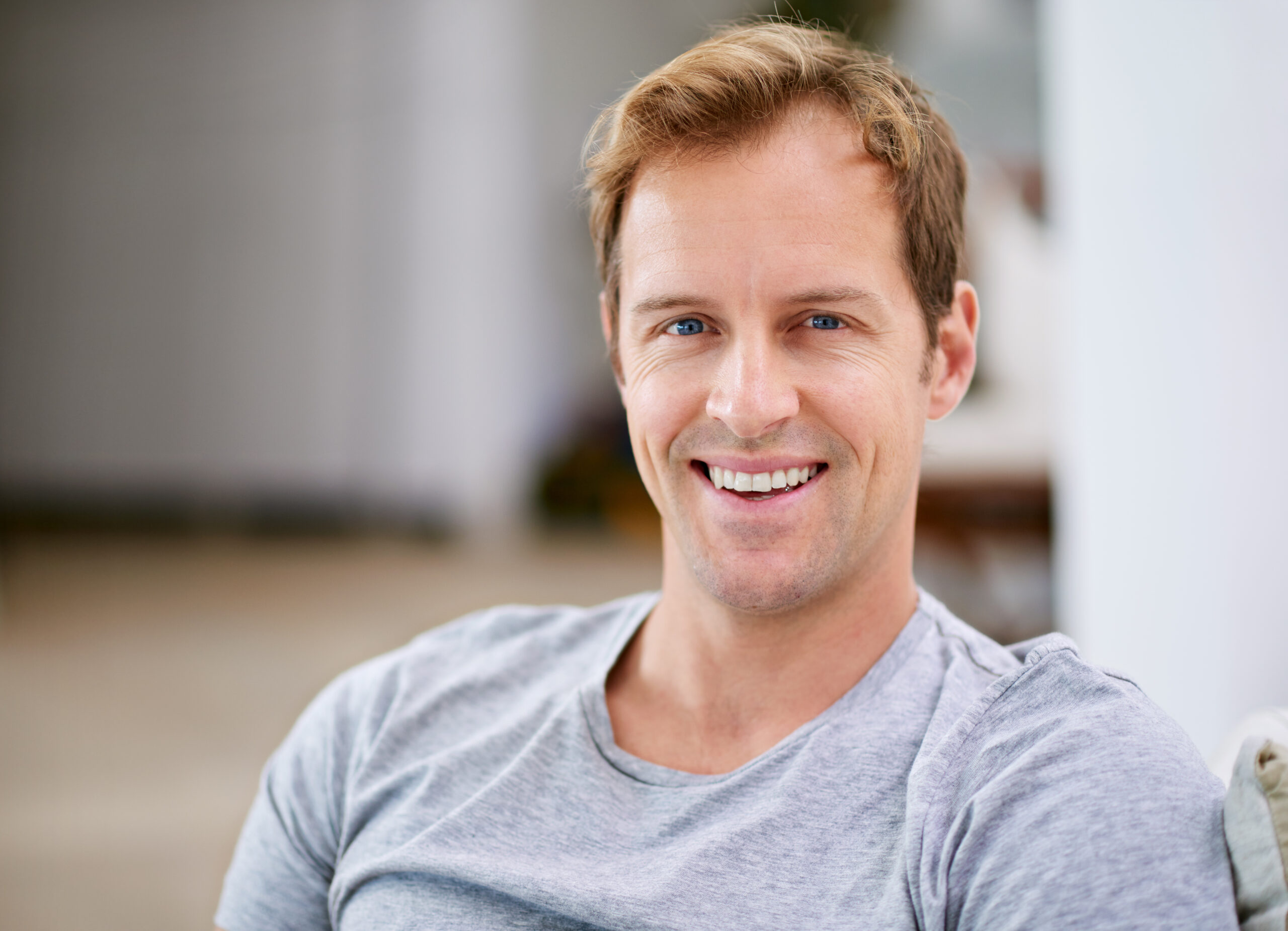 Smiling man in gray shirt against blurred indoor background at dental office.