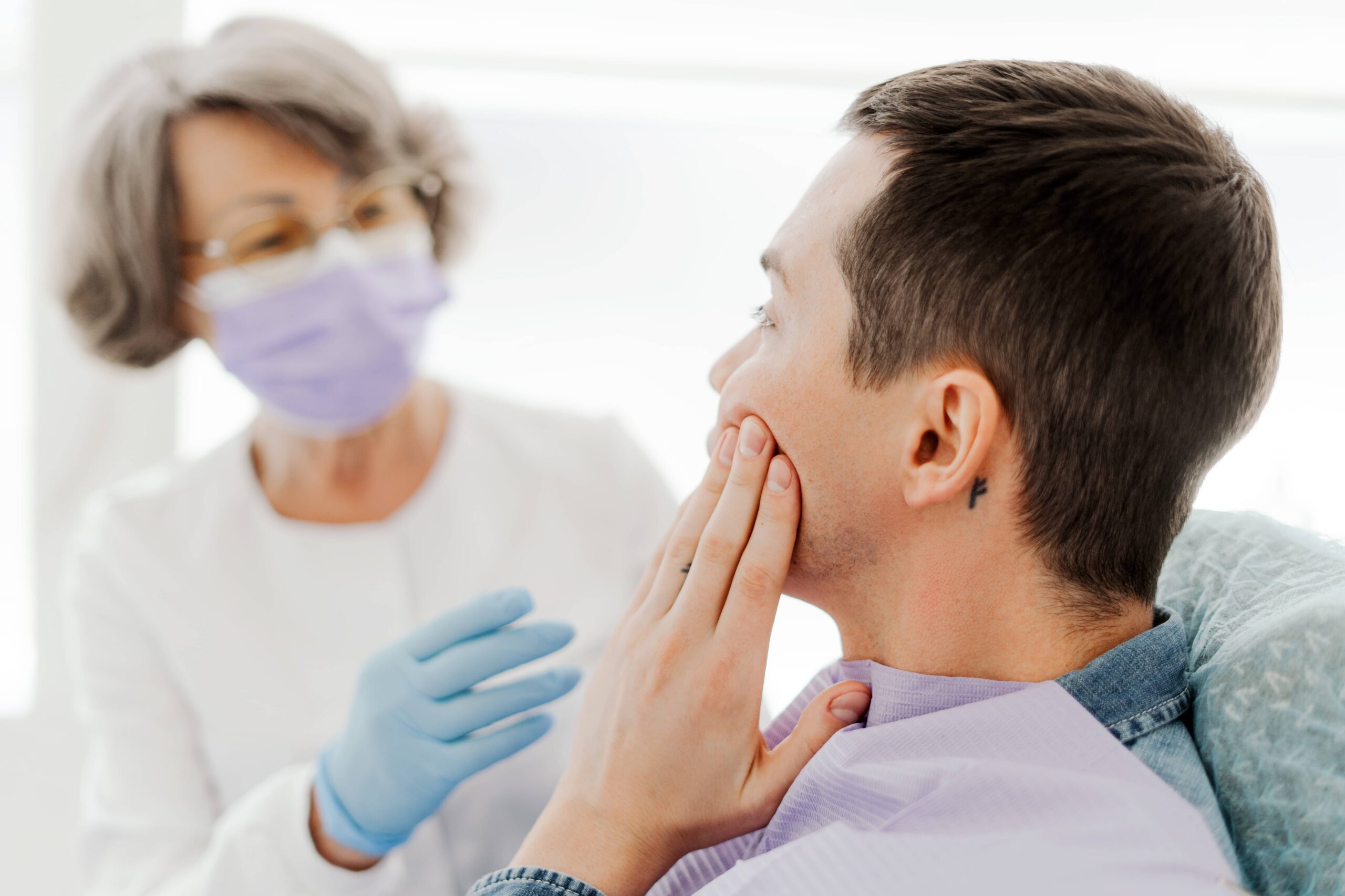 Patient holding jaw in pain while dentist examines him during emergency dental visit.